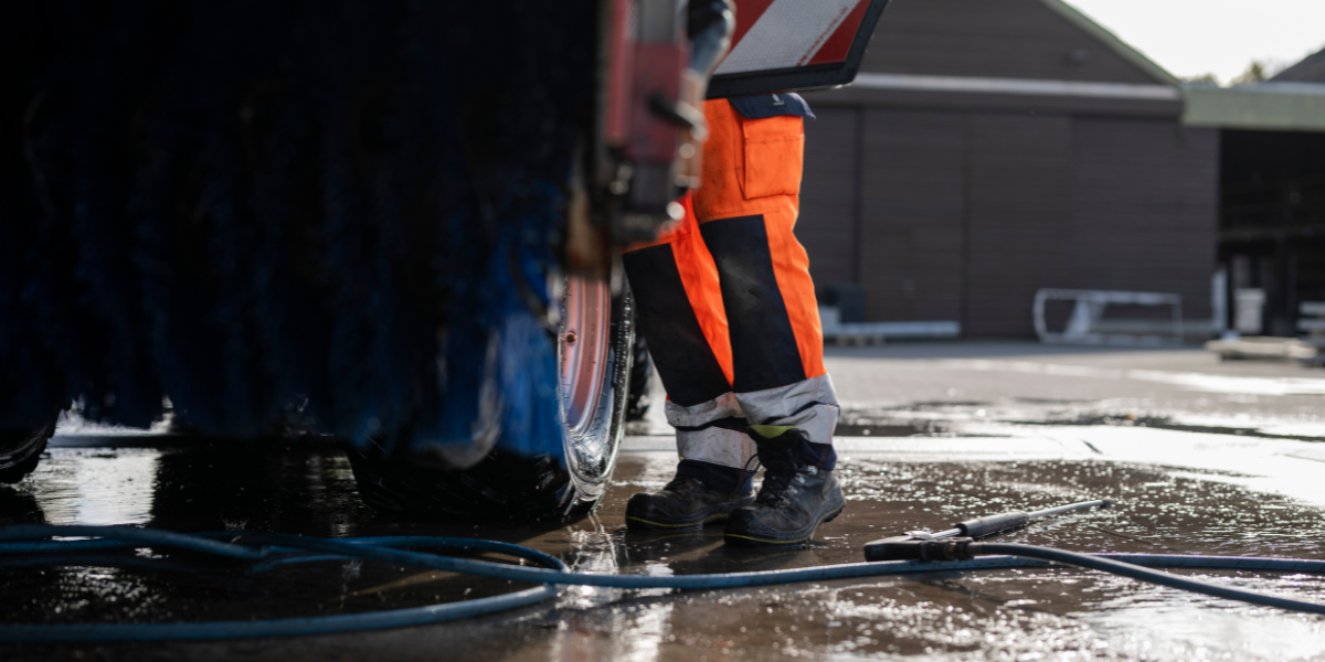 Fahrzeug in Pfütze und Wasserschlauch, daneben lehnt ein Mensch in Arbeitskleidung.